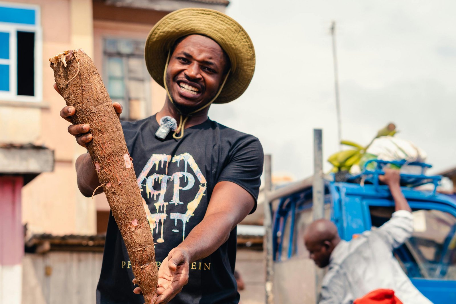 Smiling farmer proudly displays a large cassava root in an outdoor market setting.